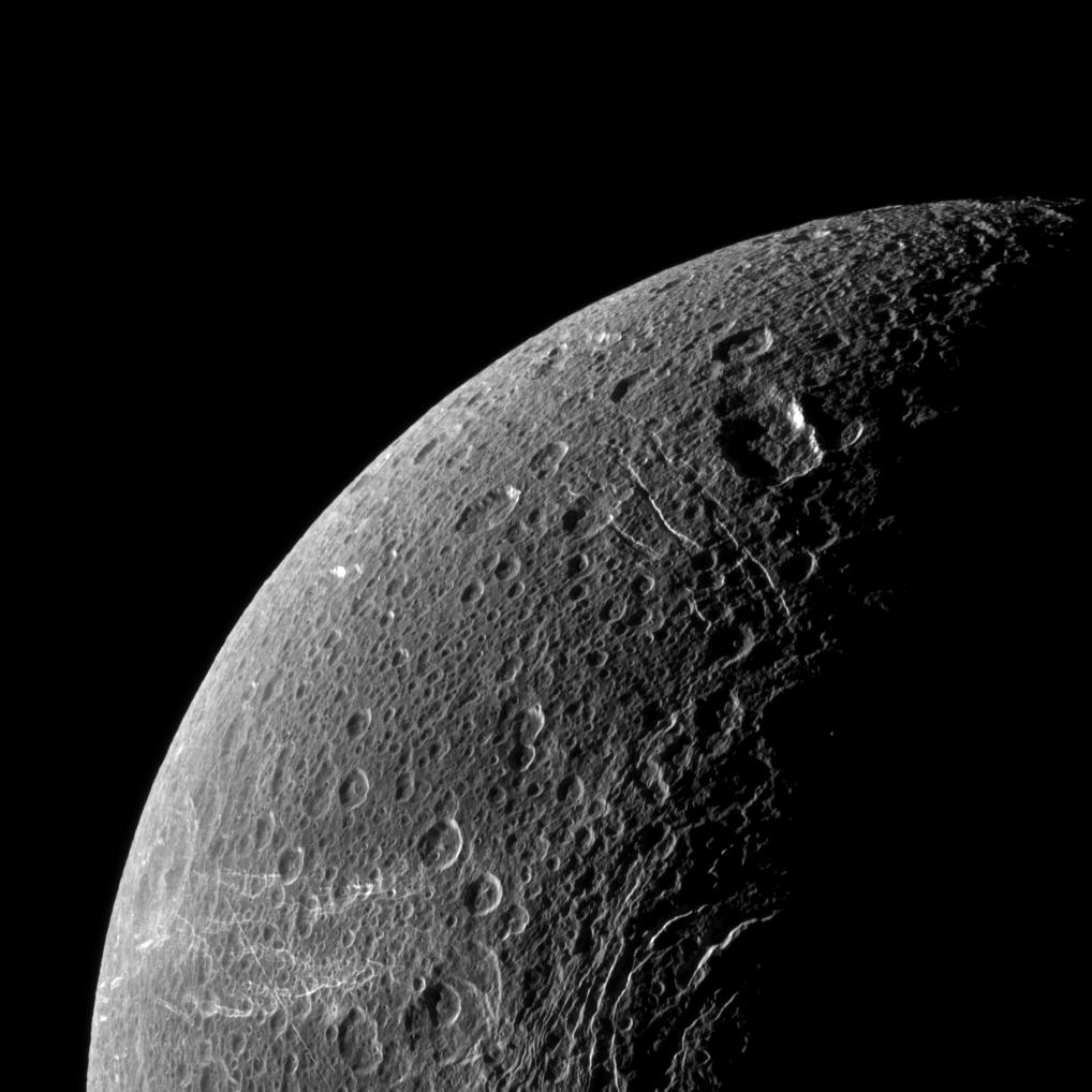 Một phần của Dione cho thấy bề mặt xám đục và những đường thì thầm màu trắng trên lọ thuốc phía dưới bên trái của hình ảnh.